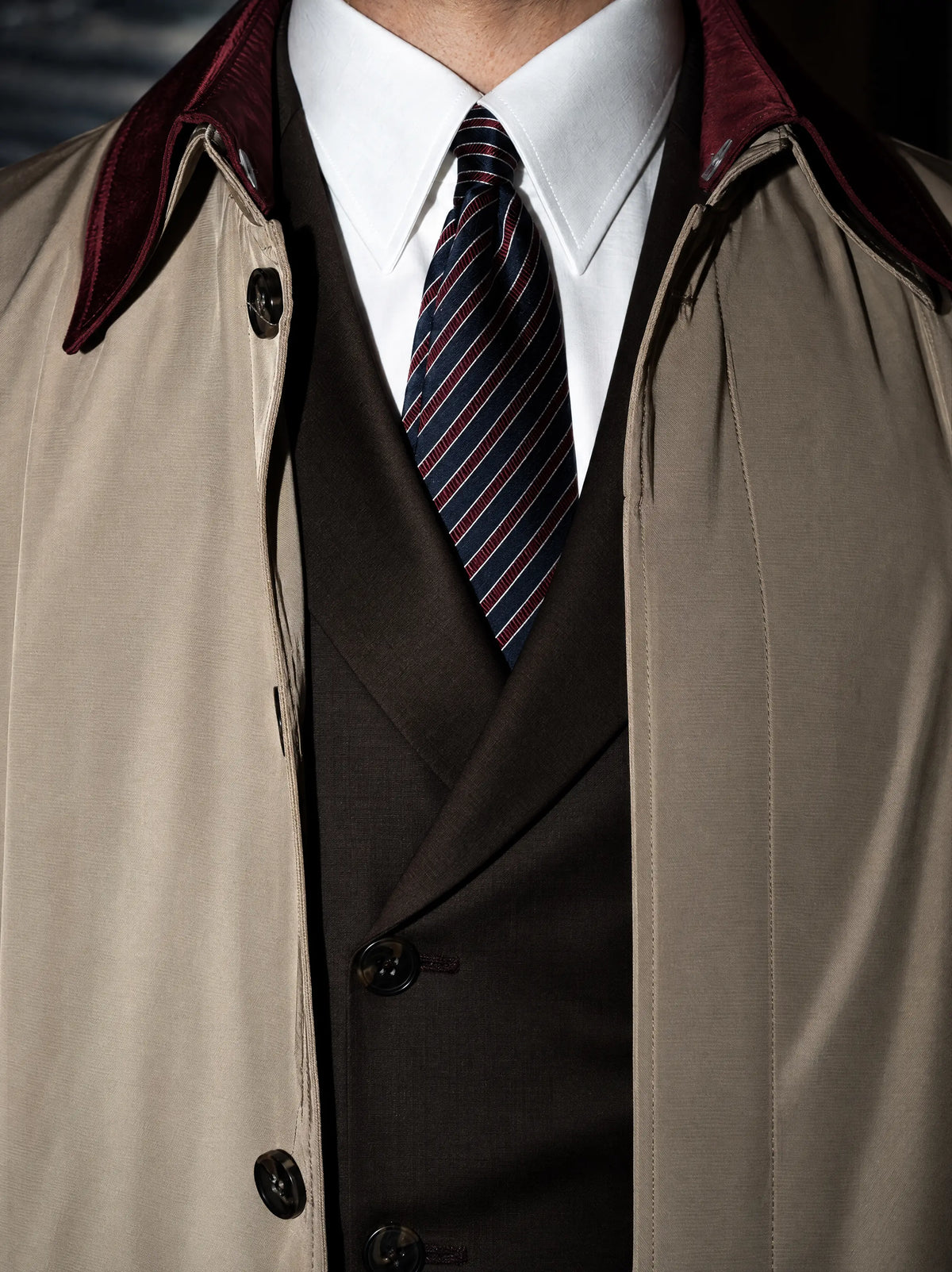 Bordo yakalı trenchcoat; altında kruvaze takım, beyaz gömlek ve çizgili kravat
